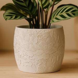 Puede incluir: Maceta de cerámica blanca con un diseño de flor de peonía en relieve. La maceta contiene una planta verde con hojas rayadas. La maceta está sobre una superficie de madera.