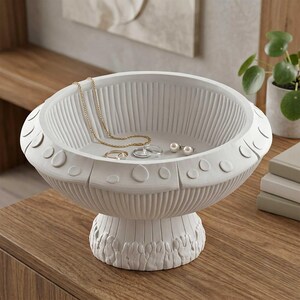 May include: A matte white pedestal bowl featuring a fluted interior, a wide rim decorated with raised circular accents, and a textured base. Inside the bowl rests a thin gold chain necklace, a simple gold ring, and two small white pearl stud earrings.