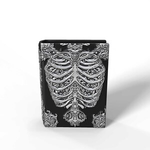 May include: A black box with a detailed silver-colored rib cage design on the front. The sides and top of the box feature ornate, swirling patterns. The overall aesthetic is gothic and decorative.