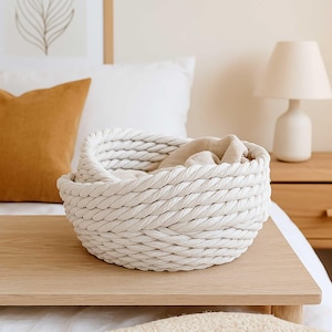 May include: A white, woven rope basket with a circular design, resting on a light wood surface. The basket contains a folded beige blanket. The background includes a white pillow, a mustard-colored pillow, and a white lamp.