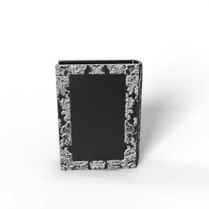 May include: A black book-shaped box with ornate silver-colored floral detailing around a central black panel. The box has a curved spine and a rectangular shape, suitable for storage or display.