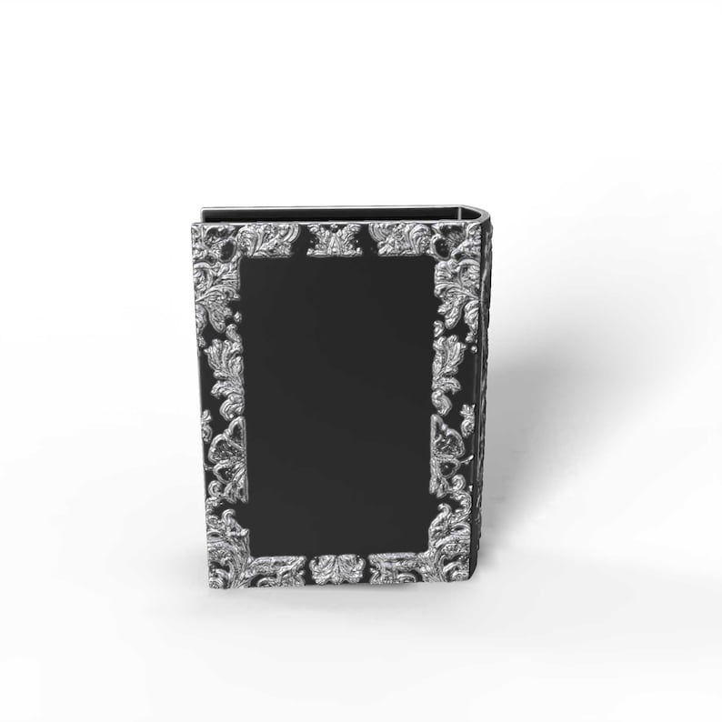 May include: A black book-shaped box with ornate silver-colored floral detailing around a central black panel. The box has a curved spine and a rectangular shape, suitable for storage or display.