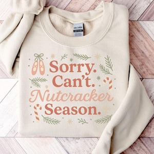 May include: A cream-coloured sweatshirt featuring the text "Sorry, Can't. Nutcracker Season." in a festive design. The design includes ballet shoes, candy canes, and sprigs of greenery. The sweatshirt is folded on a wooden surface.