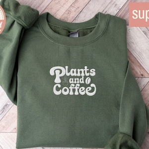 May include: A military green sweatshirt with the words "Plants and Coffee" in white script. The sweatshirt has a crew neck and long sleeves. The word "Embroidery" is in a circular logo in the upper left corner, and "super soft" is in a pink rectangle.