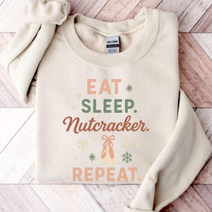 May include: A cream-coloured sweatshirt featuring the words "EAT. SLEEP. Nutcracker. REPEAT." in various colours. The design includes snowflakes and ballet shoes. A comfortable top for the festive season.