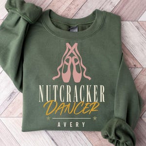 May include: An olive green crewneck sweatshirt featuring a graphic of pink ballet slippers with ribbons. The text reads NUTCRACKER DANCER in cream and gold, with the name AVERY printed below in white, accented by two small stars on a horizontal line.
