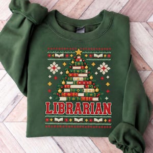 May include: A forest green crewneck sweatshirt featuring a graphic of a Christmas tree constructed from stacked books. The design includes the word LIBRARIAN in red letters, surrounded by white and yellow snowflake and book patterns.