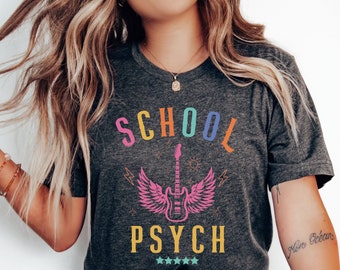 May include: A dark grey t-shirt with the text "SCHOOL PSYCH" in rainbow colors. A pink guitar with wings is in the center of the shirt. The shirt is being worn by a person with long blonde hair.