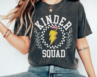 May include: A charcoal grey t-shirt with a white graphic that says "Kinder Squad" with a yellow lightning bolt inside a checkered circle. The lightning bolt has a pink stripe and glitter accents.