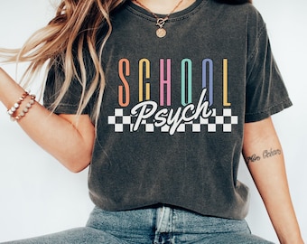 May include: Dark gray t-shirt with the words "SCHOOL Psych" in colorful, retro-style lettering. A black and white checkered pattern is below the text. The shirt is worn with blue jeans and white sneakers.