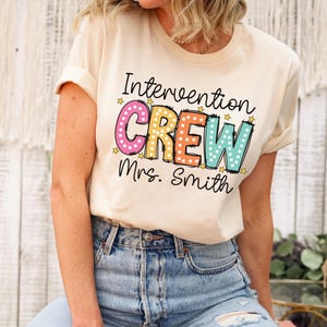 May include: Cream-colored t-shirt with the words "Intervention CREW Mrs. Smith" in a colorful, dotted design. The shirt has rolled-up sleeves and is paired with blue jeans.