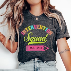 May include: A dark grey t-shirt with the text "Intervention Squad" in a rainbow arc above a pink pencil with the text "MRS. SMITH" on it.