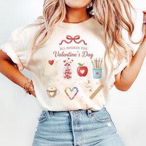 May include: Cream-colored t-shirt with the text "ALL BOOKED FOR Valentine's Day" and various Valentine's Day themed graphics. The graphics include hearts, a coffee cup, an apple, pencils, and a jar of hearts.