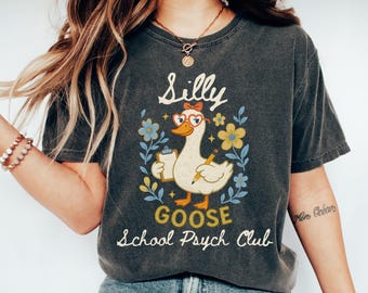 May include: Dark gray t-shirt with a whimsical graphic of a goose wearing glasses and a bow, holding a clipboard and pencil. The text reads "Silly Goose School Psych Club" in a playful font, surrounded by floral elements.