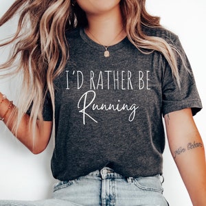 May include: A dark gray t-shirt with the text "I'd rather be running" in white lettering. The shirt is being worn by a person with long blonde hair and blue jeans.