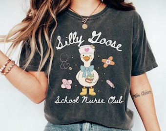 May include: Dark gray t-shirt with the words "Silly Goose School Nurse Club" and a cartoon goose wearing a nurse's hat. The goose has a blue bow tie and is holding books. Bandages, a stethoscope, and a syringe are also on the shirt.