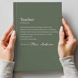 May include: A sage green notebook with the definition of "Teacher" printed in white. The definition includes the phrase "Mrs. Anderson" at the bottom. The notebook is held by two hands.