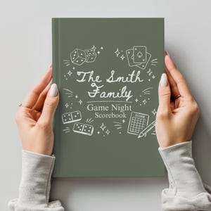 May include: A sage green game night scoreboard held by hands. The cover features white illustrations of dice, playing cards, and dominoes, with the text "The Smith Family Game Night Scorebook."