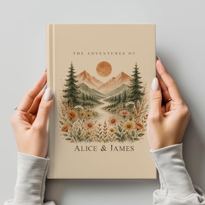 May include: A beige journal with the text "THE ADVENTURES OF" and "ALICE & JAMES." The cover features a watercolor illustration of a mountain landscape with a sun, trees, and flowers. The journal is held by two hands.