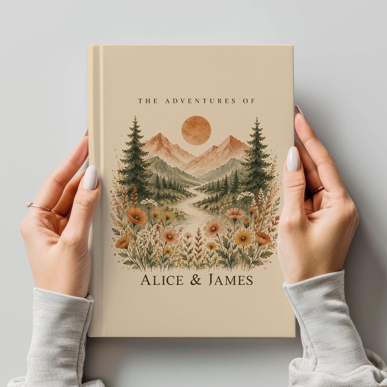 May include: A beige journal with the text "THE ADVENTURES OF" and "ALICE & JAMES." The cover features a watercolor illustration of a mountain landscape with a sun, trees, and flowers. The journal is held by two hands.