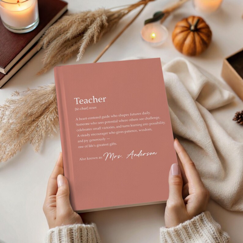 May include: A dusty rose-colored notebook with the definition of "Teacher" printed in white. The definition includes the phrase "Also known as Mrs. Anderson". The notebook is held open, with candles and a small pumpkin in the background.