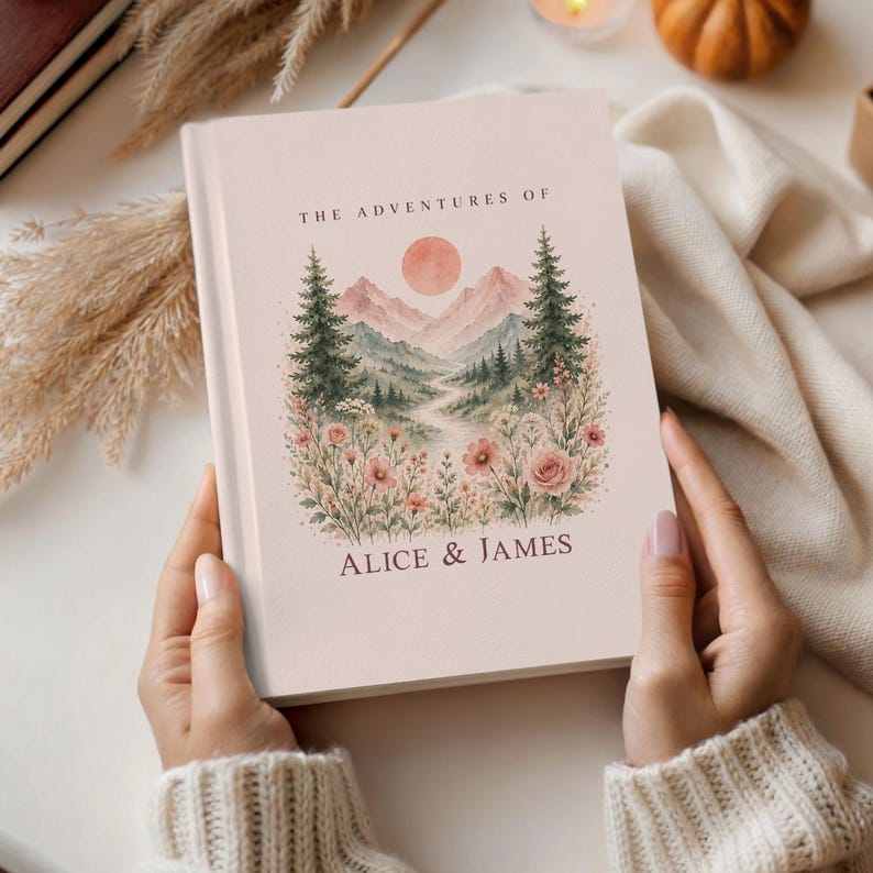 May include: A personalized journal with a soft pink cover. The cover features a watercolor landscape with mountains, trees, and flowers, and the text "THE ADVENTURES OF ALICE & JAMES".