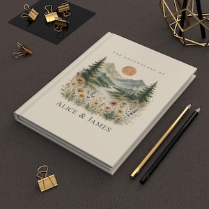 May include: A cream-colored journal with the text "THE ADVENTURES OF ALICE & JAMES" and a watercolor landscape illustration of mountains, trees, and flowers. Gold and black pencils and gold binder clips are also visible.