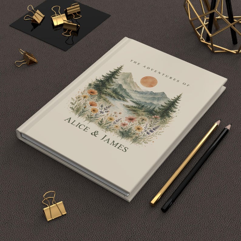 May include: A cream-colored journal with the text "THE ADVENTURES OF ALICE & JAMES" and a watercolor landscape illustration of mountains, trees, and flowers. Gold and black pencils and gold binder clips are also visible.