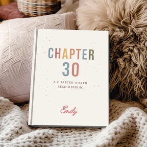 May include: A hardcover book with the text "CHAPTER 30" in colorful letters. Below, it reads "A CHAPTER WORTH REMEMBERING" and "Emily" in a cursive font. The book is set against a backdrop of soft, textured fabrics.