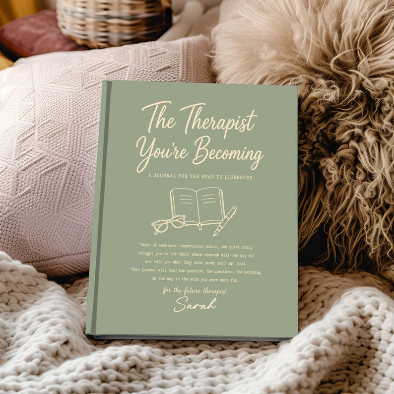 May include: A sage green hardcover journal titled The Therapist You're Becoming: A Journal for the Road to Licensure. The cover features a line drawing of an open book, glasses, and a pen, with printed text and the name Sarah at the bottom.