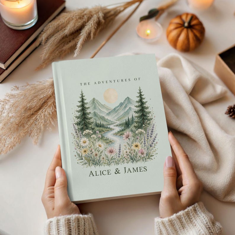 May include: A light green hardcover journal titled "The Adventures of Alice & James." The cover features a watercolor illustration of a mountain landscape with trees, flowers, and a river. The journal is held by a person.