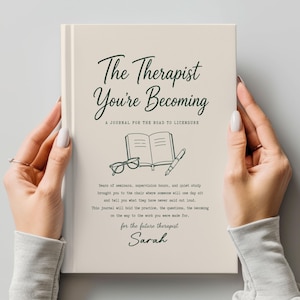 May include: A cream-colored hardcover journal titled The Therapist You're Becoming: A Journal for the Road to Licensure. The cover features dark green text, a line drawing of an open book, glasses, and a pen, with additional text and the name Sarah.