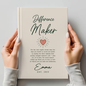 May include: A cream-colored hardcover book with dark green text reading Difference Maker, a pink heart icon, and a paragraph of text. Below is the name Emma and the text EST. 2019. The book cover features a clean, minimalist design layout.