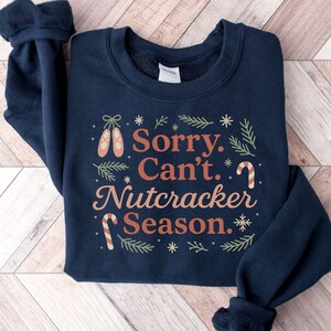 Pu&ograve; includere: Felpa blu navy con la scritta "Sorry, Can't, Nutcracker Season". Il design include scarpette da ballo, bastoncini di zucchero e fiocchi di neve nei toni del marrone e del verde. Un capo festivo e confortevole.