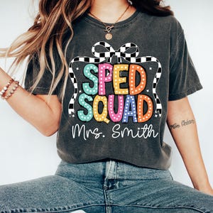 May include: Dark gray t-shirt with the text "SPED SQUAD" in colorful bubble letters, accented with a checkered bow design. Below the text is "Mrs. Smith" in a handwritten font. The shirt is a casual style.
