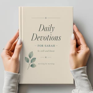 May include: A cream-coloured journal titled "Daily Devotions" with the text "For Sarah" and "be still and know." The cover features a watercolour-style leaf design. The journal is held by two hands.