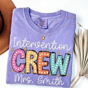 May include: A light purple t-shirt with the words "Intervention CREW Mrs. Smith" in a colorful, playful font. The word "CREW" is in pink, orange, and teal with white polka dots. A gold necklace rests on the shirt.