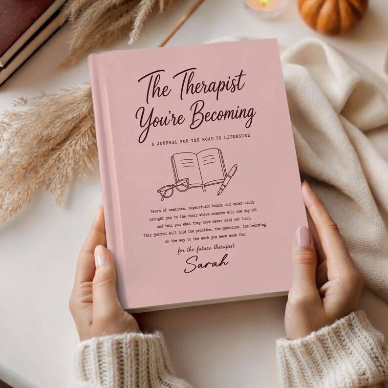 May include: A light pink journal titled The Therapist You're Becoming, A Journal for the Road to Licensure. The cover features a line drawing of an open book, glasses, and a pen, with printed text and the name Sarah at the bottom.