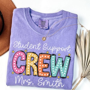 May include: A light purple t-shirt with the words "Student Support CREW Mrs. Smith" in colorful, playful lettering. The shirt has a crew neck and short sleeves. A gold necklace with a pendant is draped across the shirt.