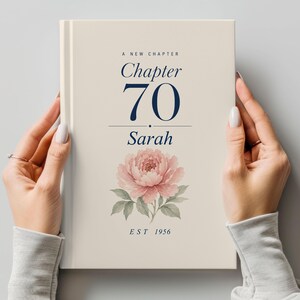 May include: A cream-coloured book with the text "A New Chapter Chapter 70 Sarah EST 1956" in navy blue. A pink peony flower with green leaves is at the bottom. The book is held by two hands.