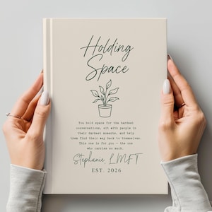 Może przedstawiać: Kremowy notatnik z napisem "Holding Space" elegancką czcionką. Poniżej ilustracja rośliny doniczkowej i tekst o wspieraniu. Notatnik jest spersonalizowany z imieniem i rokiem 2026.