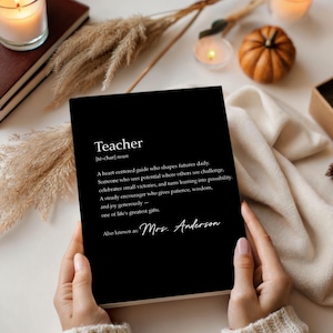 May include: A black notebook with the definition of "Teacher" printed in white. The definition includes the words "heart-centered guide," "potential," and "greatest gifts." The notebook is held by a person. The notebook is surrounded by candles and decorative items.