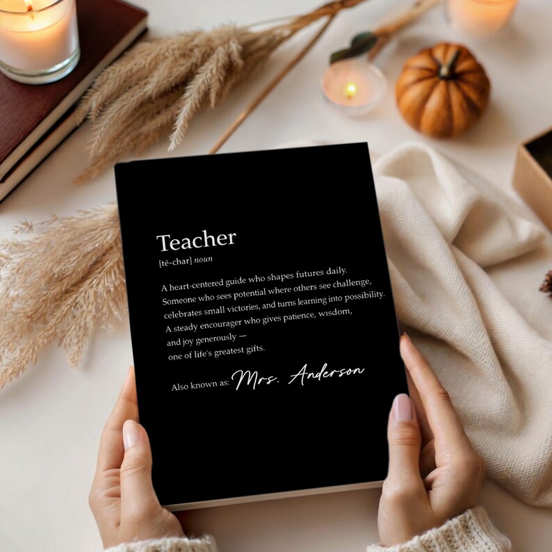 May include: A black notebook with the definition of "Teacher" printed in white. The definition includes the words "heart-centered guide," "potential," and "greatest gifts." The notebook is held by a person. The notebook is surrounded by candles and decorative items.