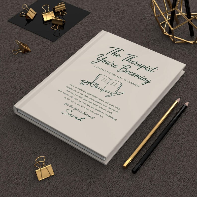 May include: A white hardcover journal titled The Therapist You're Becoming, A Journal for the Road to Licensure. The cover features a line drawing of glasses, an open book, and a pen, with a paragraph of text and the name Sarah printed below.