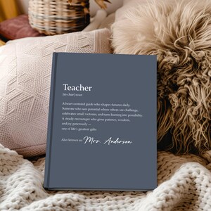 May include: A navy blue hardcover book with the definition of "Teacher" printed in white. The definition includes the phrase "Mrs. Anderson" at the bottom. The book is resting on a cream-colored knitted blanket.