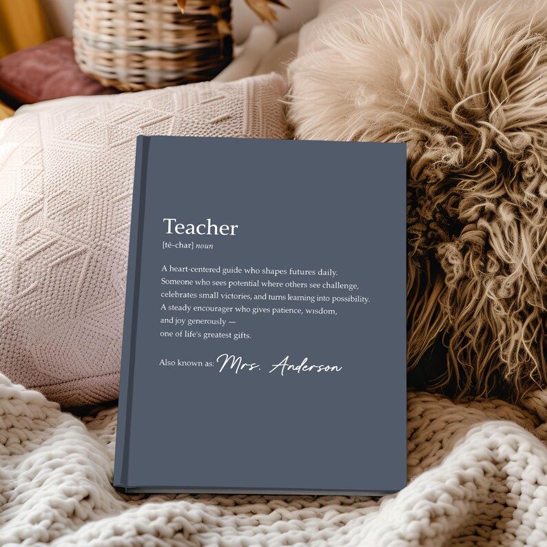 May include: A navy blue hardcover book with the definition of "Teacher" printed in white. The definition includes the phrase "Mrs. Anderson" at the bottom. The book is resting on a cream-colored knitted blanket.