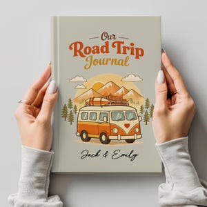 May include: A journal with the text "Our Road Trip Journal" in orange and brown. The cover features a vintage orange and white van with a heart on the front, mountains, trees, and the names "Jack & Emily" in script.
