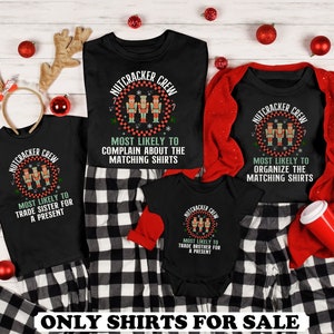 May include: A set of four black t-shirts with a Christmas Nutcracker theme. The shirts feature a graphic of three Nutcrackers and the text "Nutcracker Crew". Each shirt has a different message about the person wearing it. The shirts are on a white wooden background with red and white plaid fabric and red ornaments.