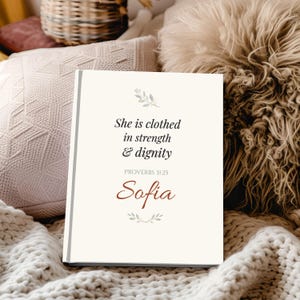 May include: A canvas print featuring the quote "She is clothed in strength & dignity" and the name "Sofia". The text is in black and brown, with leaf accents. The background is a cream colour.