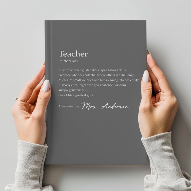 May include: A gray notebook with the definition of "Teacher" printed in white. The definition includes the phrase "Mrs. Anderson" at the bottom. The notebook is held by two hands.